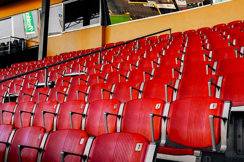 Fileiras de assentos vermelhos vazios e numerados em uma arquibancada coberta de estádio de futebol, com vista em ângulo inclinado mostrando a extensão das cadeiras